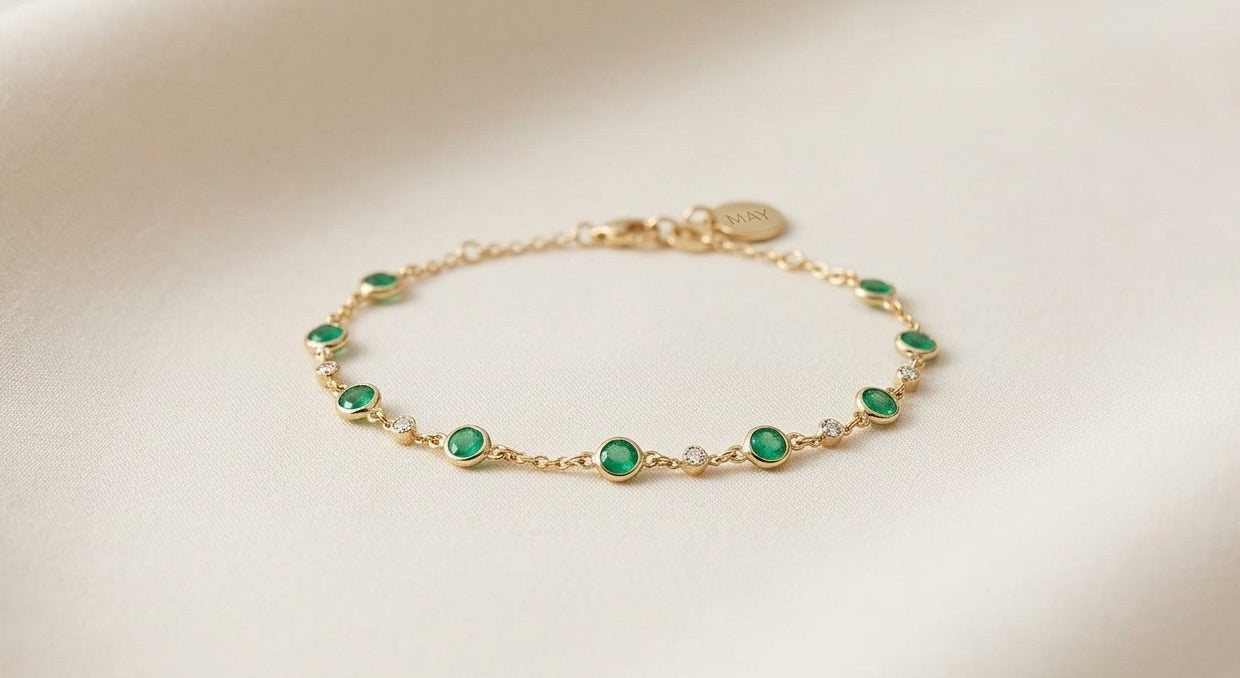 Néhlia's herinneringssieraden: Gouden geboortesteen armband met groene smaragd op een serene ondergrond.